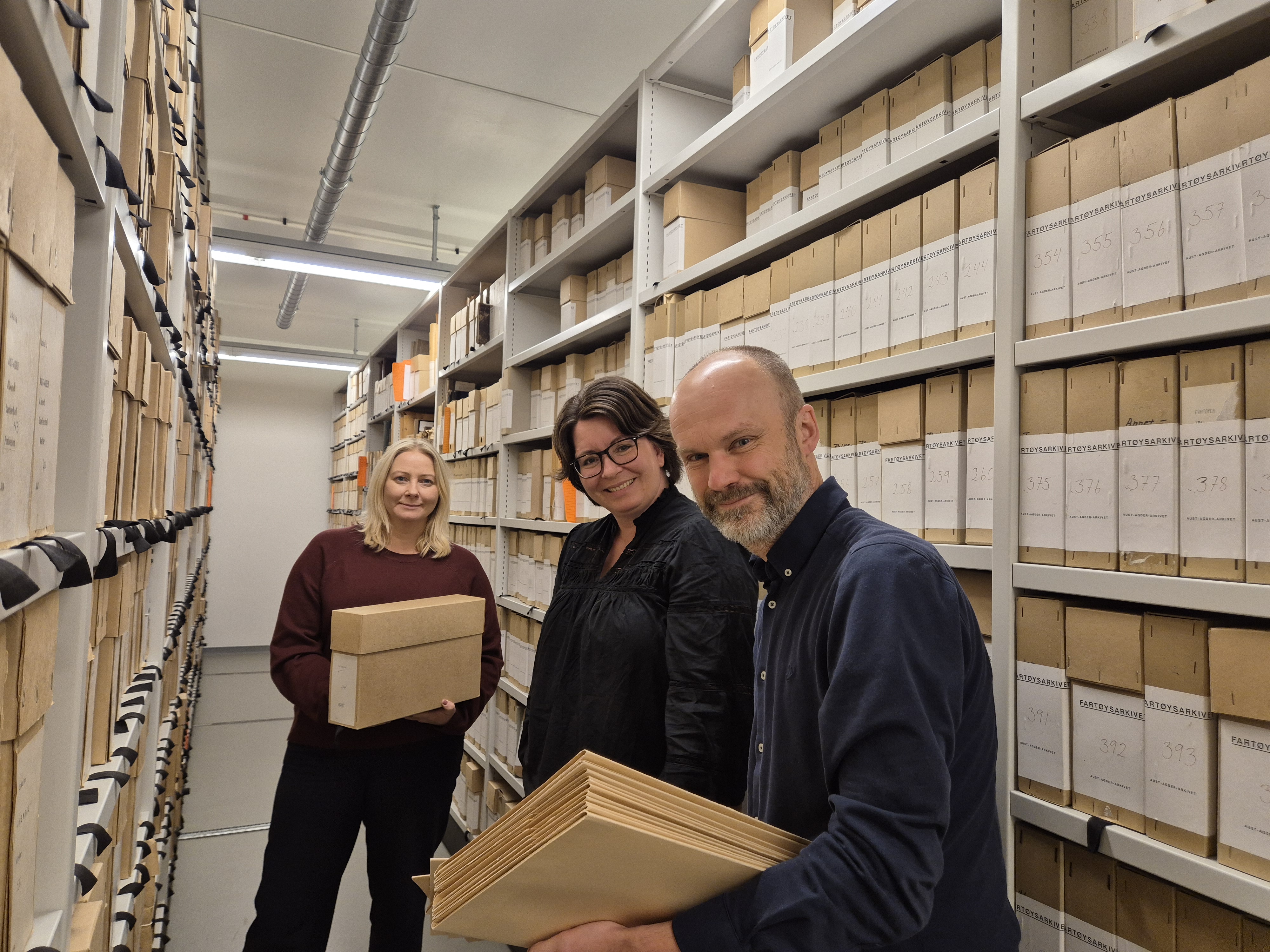 Bilde av tre personer i et arkiv, med arkivmapper i hendene