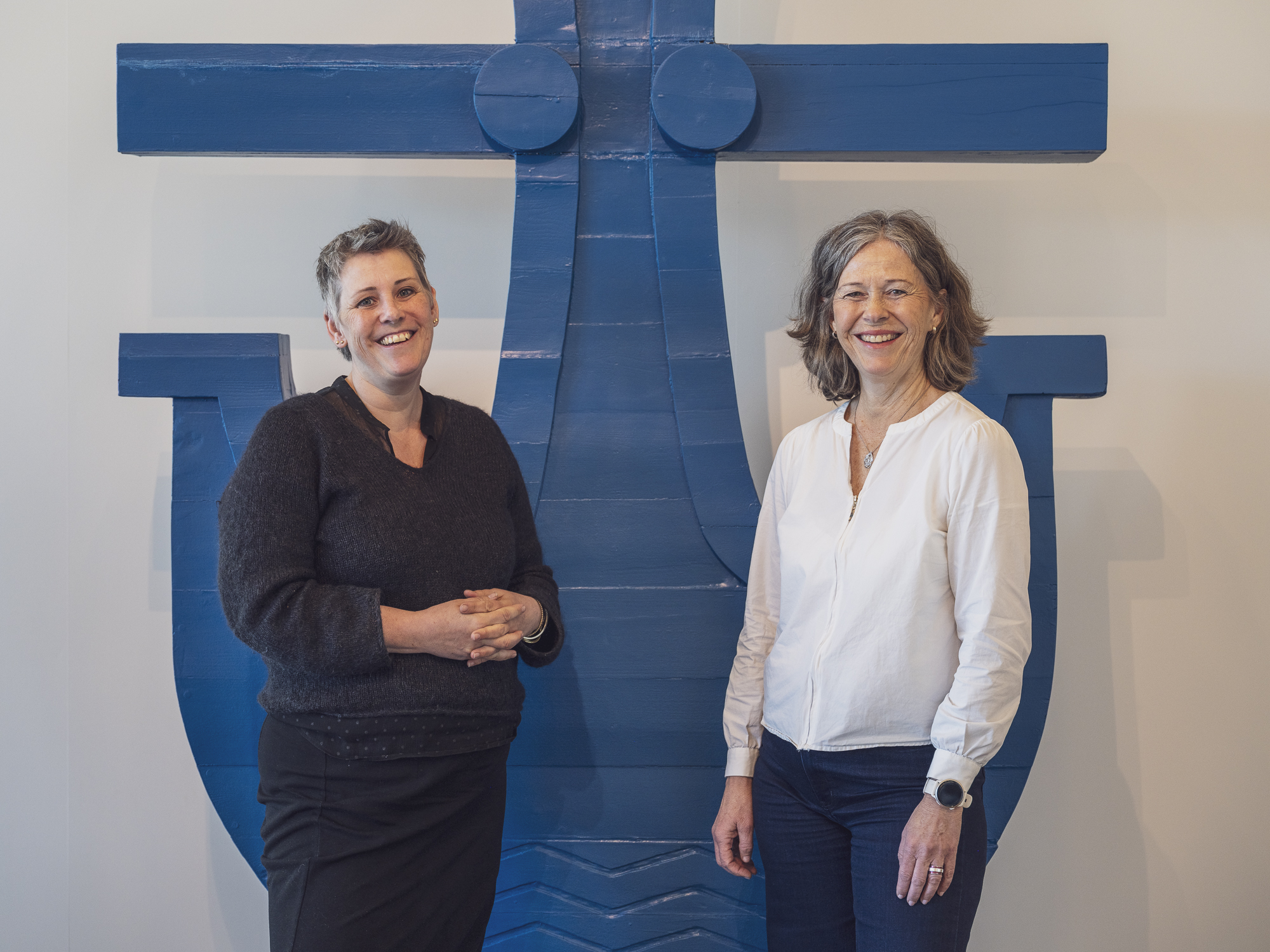 To smilende damer foran blått anker