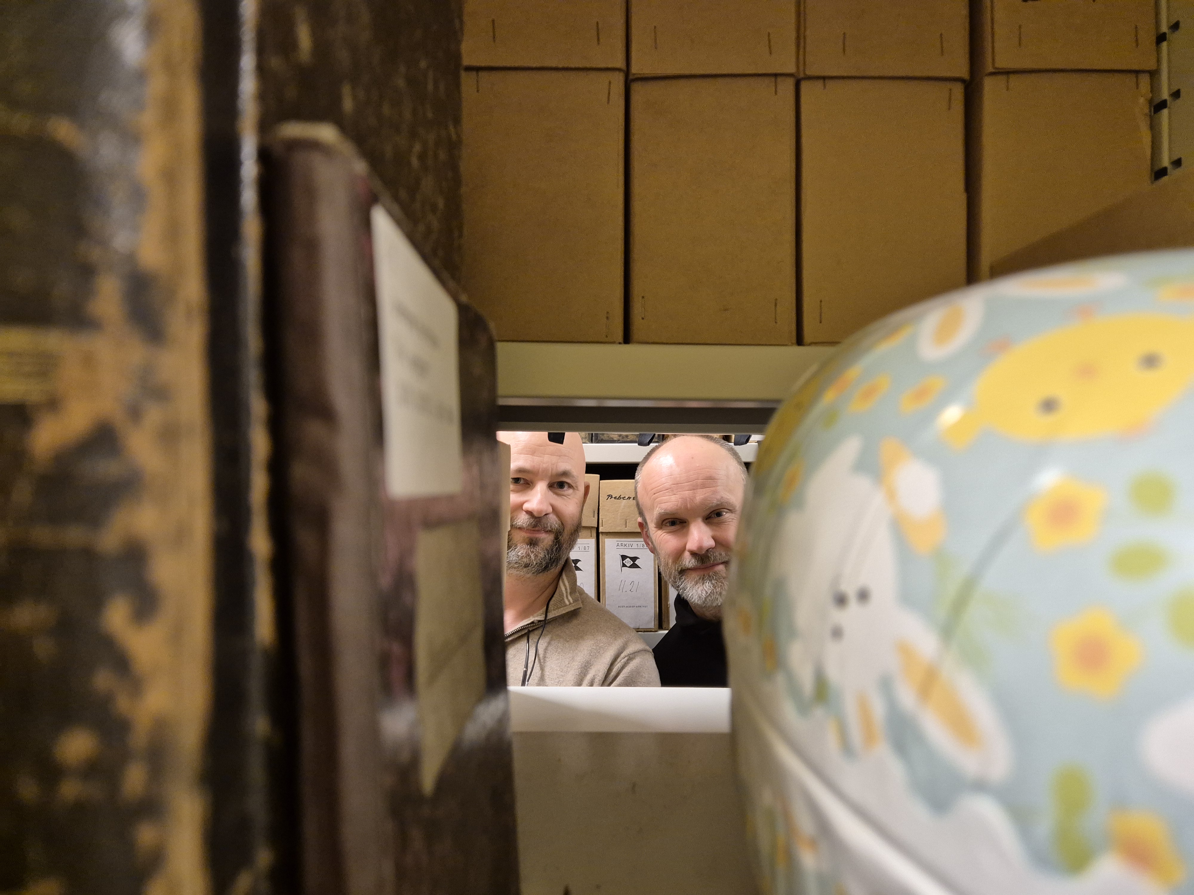 To personer titter frem gjennom en akrivhylle, gamle bpker til venstre g et påskeegg til høyre. 